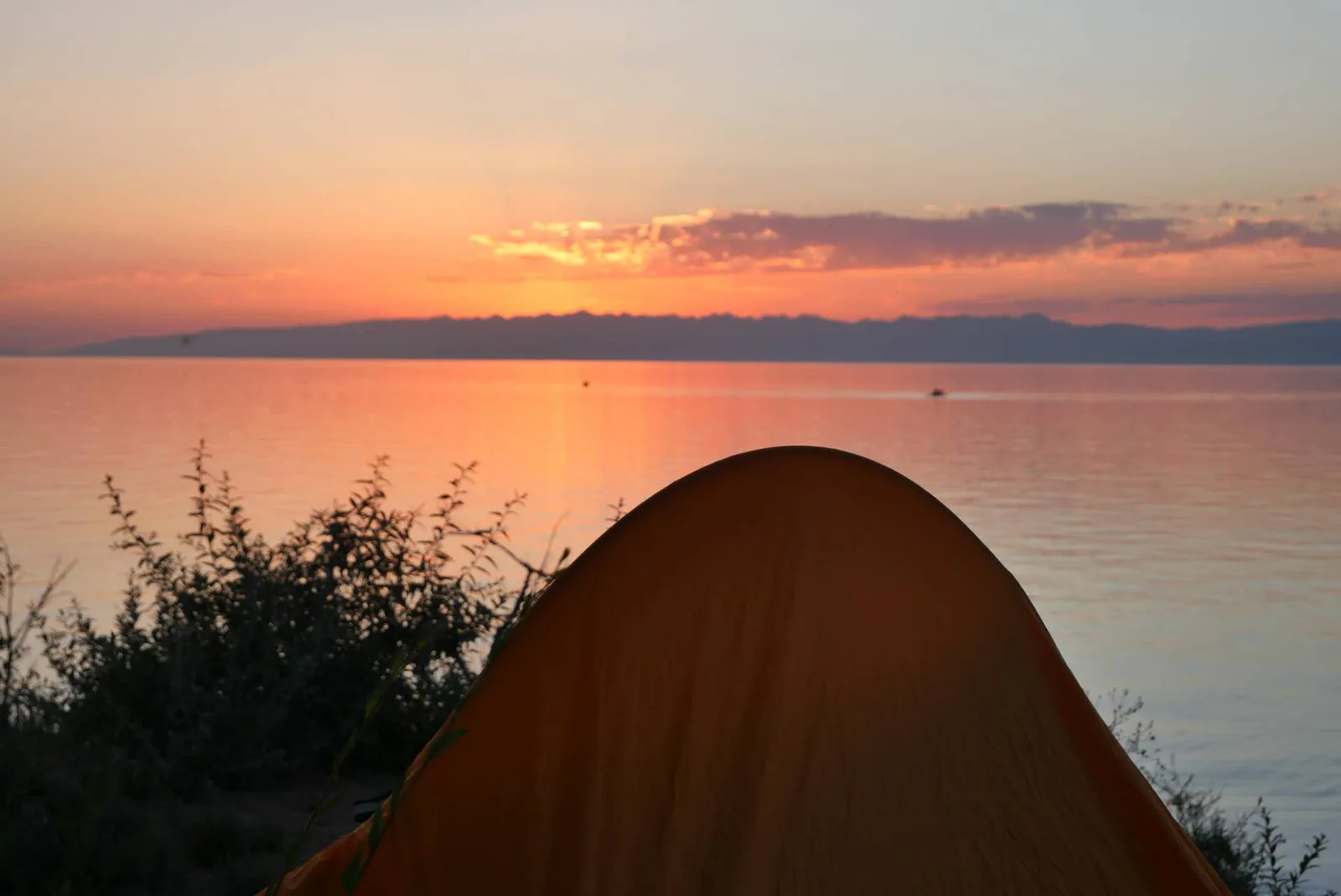 Orange tent among shrubs beside Issyk-Kul at sunset, with mountains across the lake